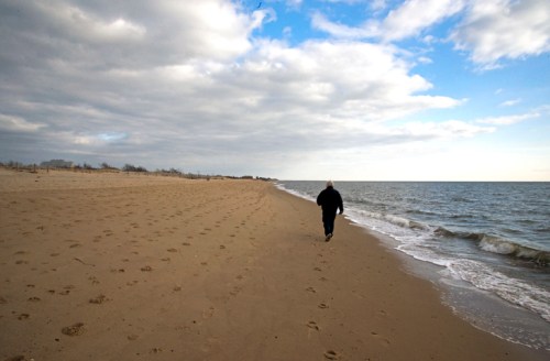A person walks along a sandy beach with gentle waves and a cloudy sky in the background.
