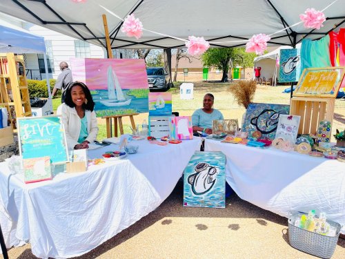 Two women smile behind a table filled with colorful art and crafts at an outdoor market under a tent.