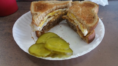 A toasted sandwich with eggs and sausage, served with sliced pickles on a paper plate.