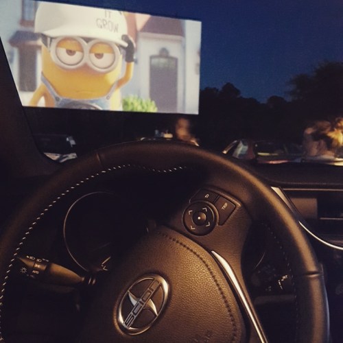 A car's steering wheel in the foreground with a drive-in movie screen showing a Minion character in the background at night.