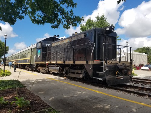 A black locomotive with a green passenger car parked at a station under a partly cloudy sky.