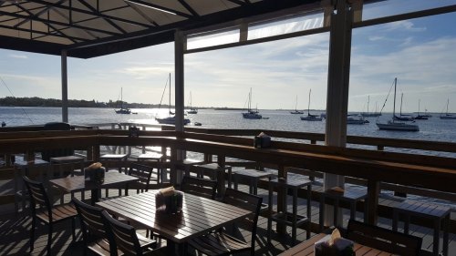A waterfront view from a restaurant deck, featuring wooden tables and sailboats on the water under a clear sky.