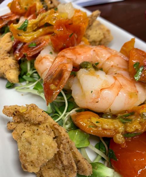 A close-up of a plate featuring shrimp, fried items, and garnished with herbs and tomatoes on a bed of microgreens.