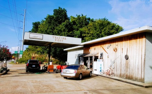 A rustic trading post with a gas station, surrounded by trees and blue sky, featuring a wooden facade and vintage decor.