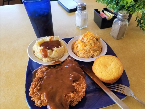 A plate of fried chicken with gravy, mashed potatoes, cornbread, and a side dish, accompanied by a drink.