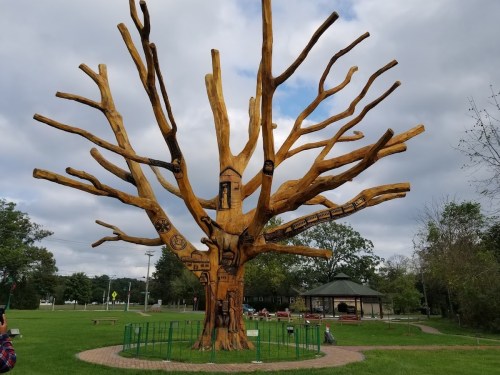A large, artistic wooden tree sculpture with intricate carvings, set in a grassy park under a cloudy sky.