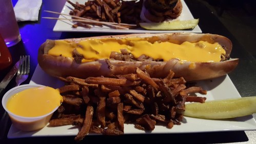 A large hot dog topped with melted cheese, served with crispy fries and a pickle on a white plate.
