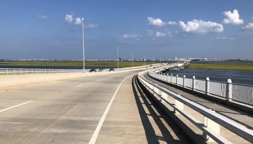 A curved bridge stretches over water, with a clear blue sky and scattered clouds in the background.