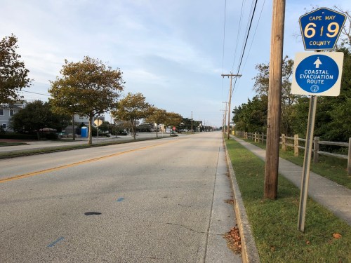 A quiet road with trees lining the sides, featuring a sign for Cape May County and a coastal evacuation route.