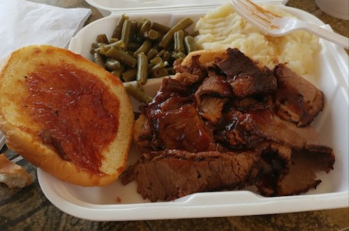 A takeout container with sliced barbecue beef, mashed potatoes, green beans, and a slice of bread with sauce.