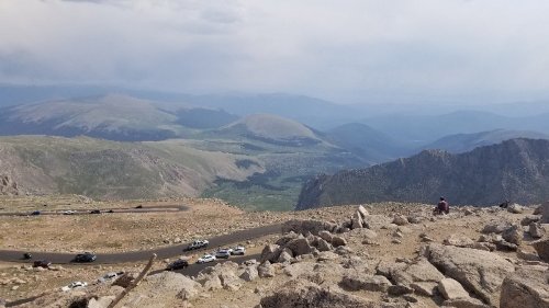 A panoramic view of mountains and valleys, with a winding road and cars visible in the foreground.