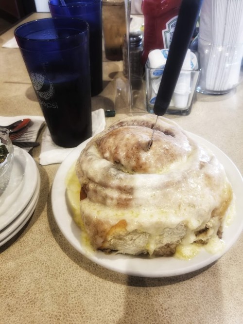 A large, gooey cinnamon roll on a plate, topped with icing, next to a glass of soda and condiments on a table.