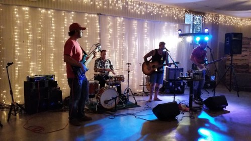 A band performs on stage with instruments, surrounded by twinkling lights in a festive setting.