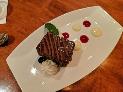 A chocolate dessert on a white plate, garnished with mint, whipped cream, and colorful sauces.