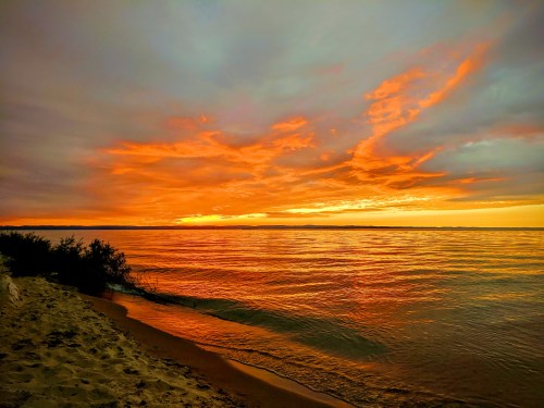 A serene beach at sunset, with vibrant orange and yellow hues reflecting on calm water under a cloudy sky.