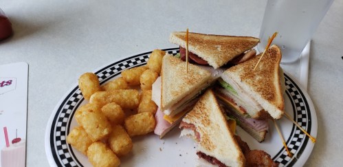 A plate with a stacked sandwich and crispy tater tots, served with a glass of water.