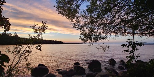 A serene lakeside view at sunset, with colorful clouds reflecting on the water and framed by trees and rocks.