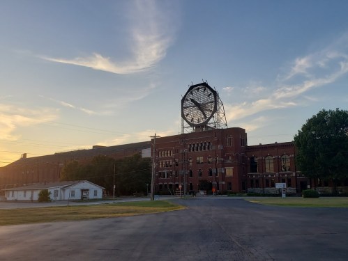 A large brick building with a circular structure on top, set against a sunset sky and grassy foreground.