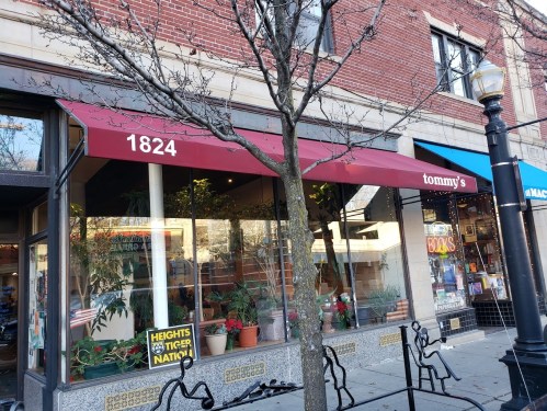 A storefront with a maroon awning labeled "tommy's," featuring plants and a sign in the window, on a brick building.