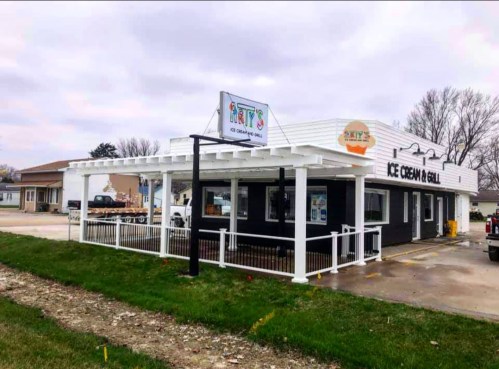 A modern ice cream shop with a white pergola and sign, surrounded by grass and a cloudy sky.