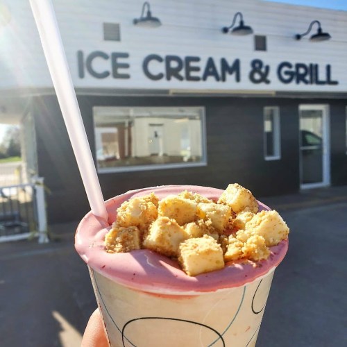 A hand holds a pink ice cream topped with cake pieces, in front of an ice cream shop with a grill sign.