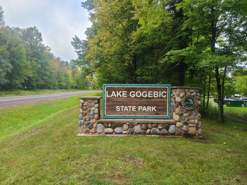Sign for Lake Gogebic State Park, surrounded by trees, with a gravel road in the background.
