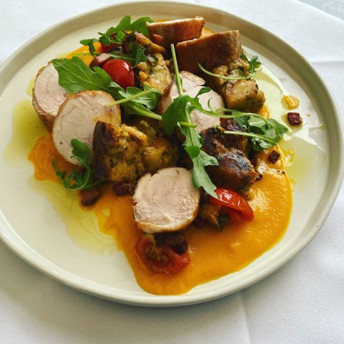 A plate of sliced chicken with stuffing, cherry tomatoes, and arugula on a bed of orange sauce, drizzled with olive oil.