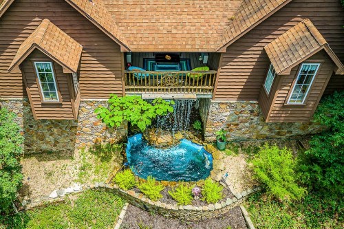 Aerial view of a brown house with a waterfall and pond in the front yard, surrounded by greenery and landscaping.