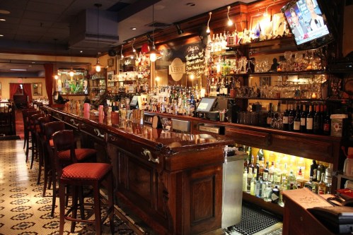 A well-stocked bar with wooden accents, shelves of bottles, and a cozy atmosphere, featuring bar stools and decorative lighting.