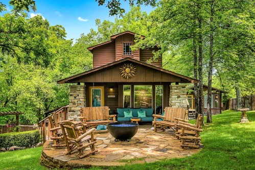 A cozy cabin surrounded by trees, featuring a stone patio with wooden chairs and a fire pit in the foreground.