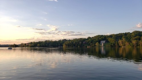 A serene lake at sunset, reflecting trees and clouds, with a few boats and houses along the shore.