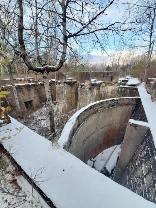Abandoned concrete structure surrounded by trees and snow, with a cloudy sky in the background.