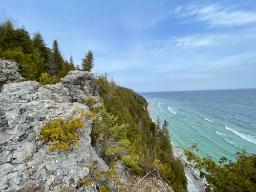 A rocky cliff overlooks a serene coastline with gentle waves and lush greenery under a clear blue sky.