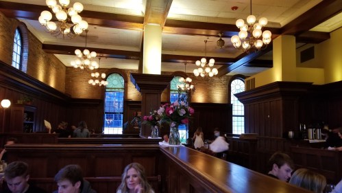 A cozy restaurant interior with wooden decor, large windows, and floral arrangements, featuring diners at tables.