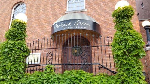 Exterior of Orchard Green restaurant, featuring a brick facade, decorative gate, and lush green vines.
