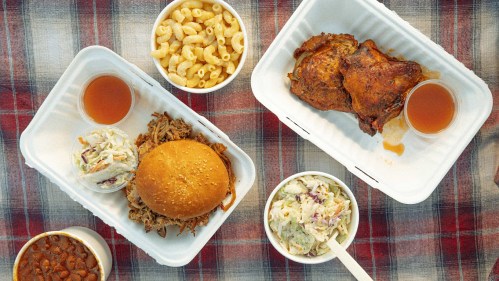 Two takeout trays with pulled pork sandwich, coleslaw, macaroni, baked beans, and a piece of grilled chicken.