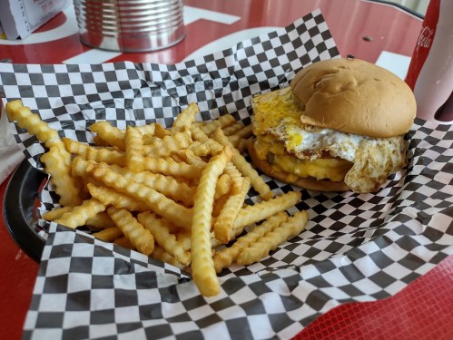 A plate with crinkle-cut fries and a burger topped with a fried egg, served in a checkered paper basket.