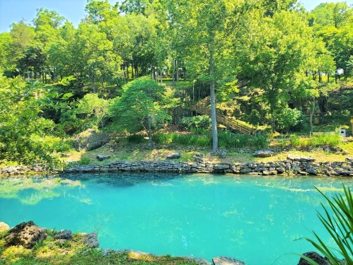 A serene turquoise pond surrounded by lush green trees and rocky banks, creating a peaceful natural landscape.