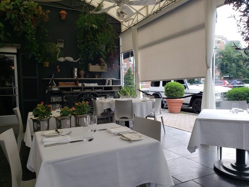 A cozy restaurant with white tablecloths, potted plants, and a partially open awning, featuring a view of parked cars outside.