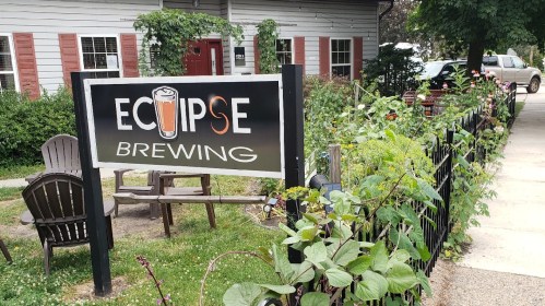Sign for Eclipse Brewing in front of a garden, with chairs and a building in the background.