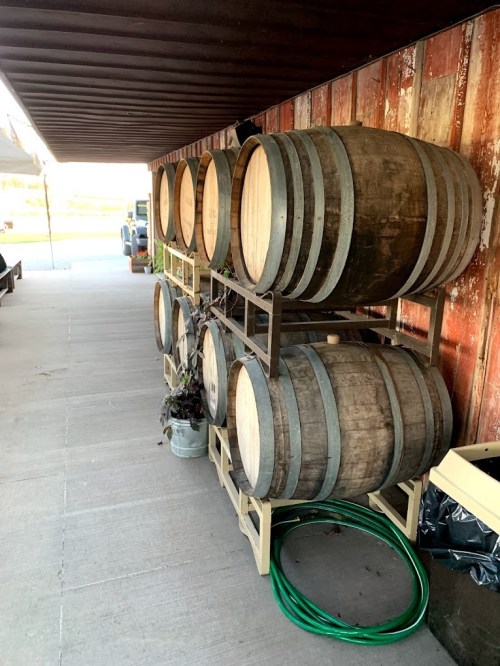 A stack of wooden barrels on a stand against a rustic red wall, with a green hose coiled nearby.