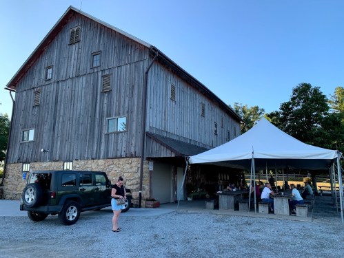 A rustic barn with a stone base and a tent outside, where people are gathered. A parked Jeep is in the foreground.