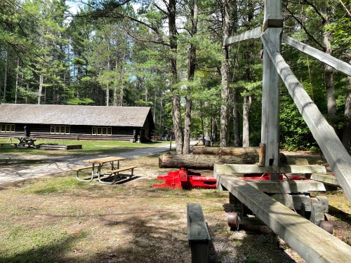 A wooden catapult stands in a forested area near a log cabin and picnic tables. Trees surround the scene.