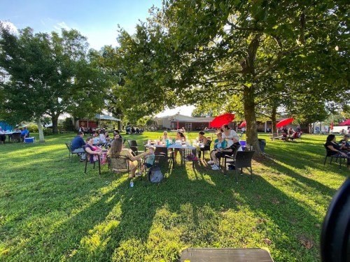 A group of people sitting around tables in a grassy area, enjoying a sunny day under trees and umbrellas.