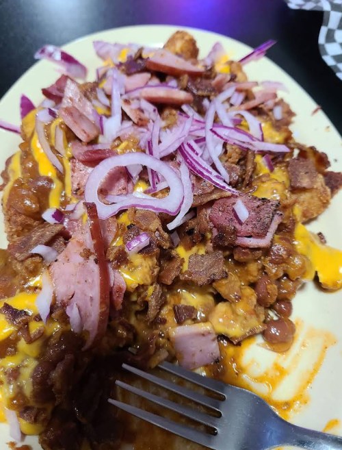 A plate of loaded nachos topped with cheese, bacon, ham, onions, and chili, served with a fork on the side.