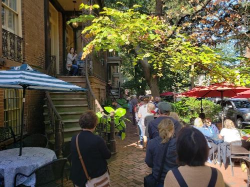A line of people waiting outside a building, with greenery and outdoor seating visible in a sunny setting.