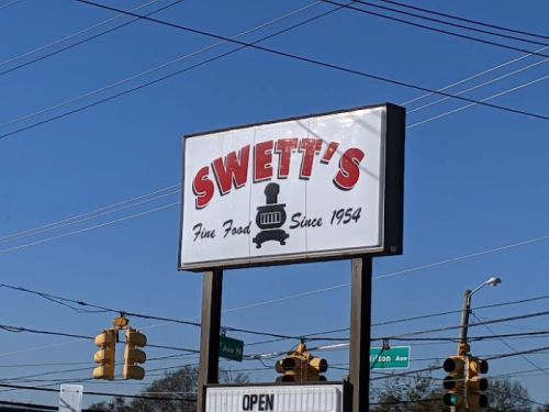 Sign for Swett's restaurant, featuring "Fine Food Since 1954" and an "Open" message, with traffic lights in the background.