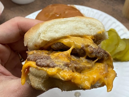 A hand holding a cheesy double burger with melted cheddar, served on a bun, with pickles in the background.