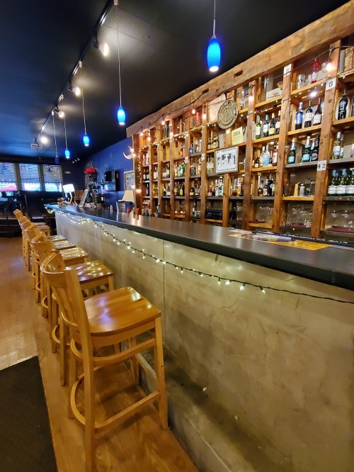 A cozy bar with wooden shelves filled with bottles, illuminated by blue pendant lights and lined with wooden stools.