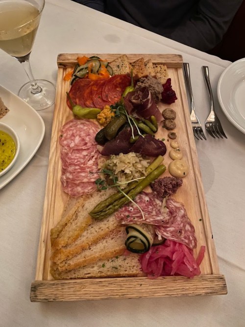 A wooden platter filled with assorted meats, pickles, bread, and garnishes, accompanied by a glass of wine.